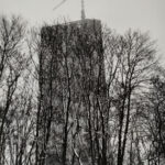 Ein hohes Gebäude im Bau mit einem Kran auf dem Dach, teilweise verdeckt von kahlen Bäumen im Vordergrund. Die Szene scheint im Winter zu spielen. (KI generierte Beschreibung)