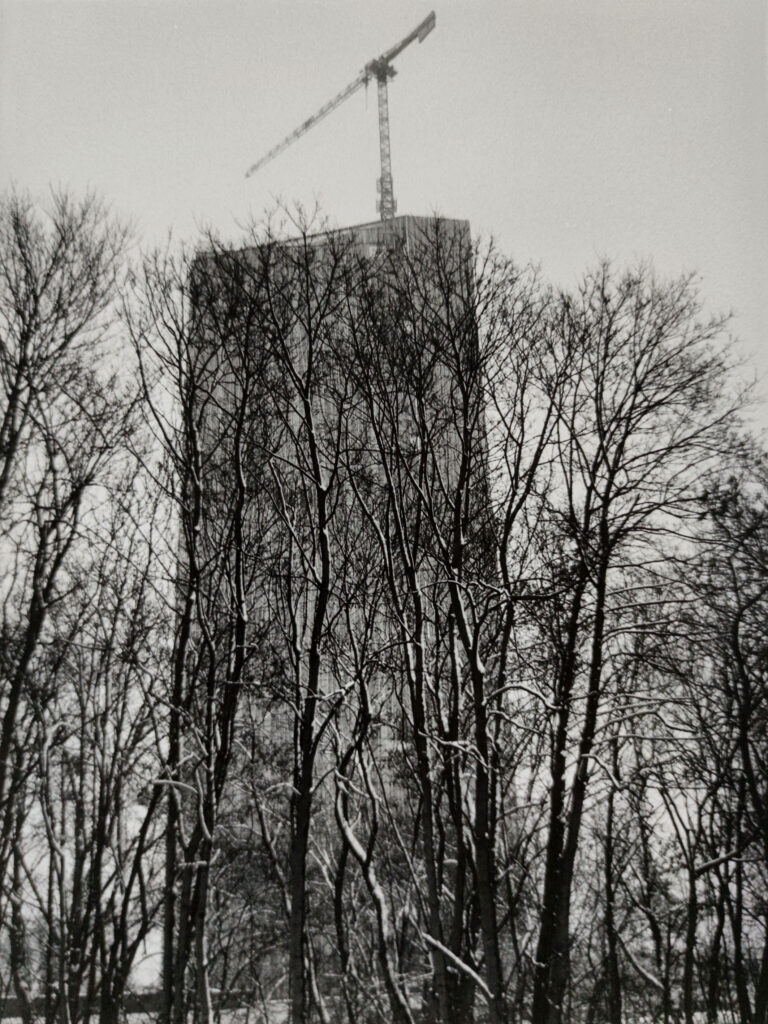 Ein hohes Gebäude im Bau mit einem Kran auf dem Dach, teilweise verdeckt von kahlen Bäumen im Vordergrund. Die Szene scheint im Winter zu spielen. (KI generierte Beschreibung)