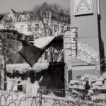 Ein Schwarz-Weiß-Foto zeigt ein Gebäude mit einem Aldi-Schild, das gerade von Baumaschinen abgerissen wird; an der noch stehenden Wand ist Graffiti zu sehen. (KI generierte Beschreibung)