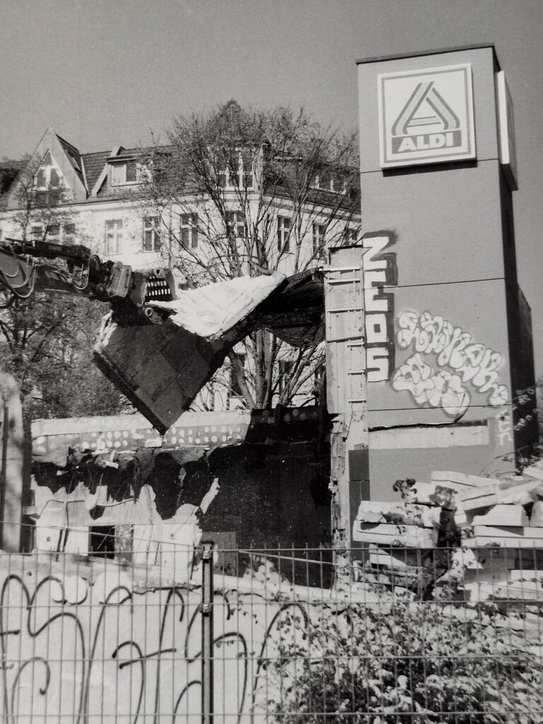 Ein Schwarz-Weiß-Foto zeigt ein Gebäude mit einem Aldi-Schild, das gerade von Baumaschinen abgerissen wird; an der noch stehenden Wand ist Graffiti zu sehen. (KI generierte Beschreibung)
