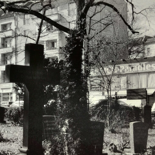 Ein Friedhof mit Grabsteinen und einem großen Kreuz im Vordergrund, im Hintergrund sind Wohnblocks und eine Werbetafel mit der Aufschrift „Paradies BERLIN“ zu sehen. (KI generierte Beschreibung)