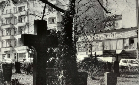 Ein Friedhof mit Grabsteinen und einem großen Kreuz im Vordergrund, im Hintergrund sind Wohnblocks und eine Werbetafel mit der Aufschrift „Paradies BERLIN“ zu sehen. (KI generierte Beschreibung)