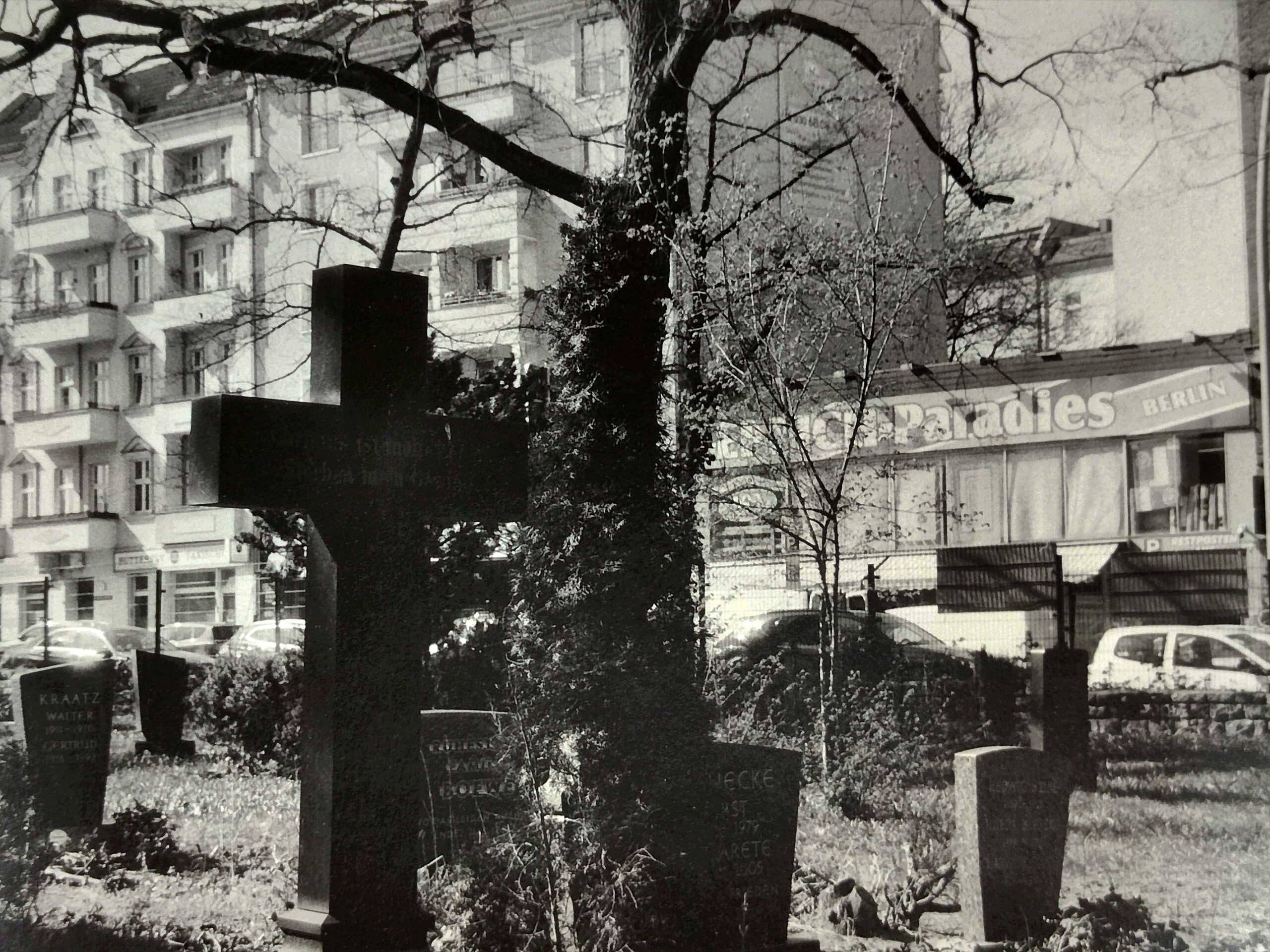 Ein Friedhof mit Grabsteinen und einem großen Kreuz im Vordergrund, im Hintergrund sind Wohnblocks und eine Werbetafel mit der Aufschrift „Paradies BERLIN“ zu sehen. (KI generierte Beschreibung)