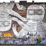 Große Wandtapete mit zwei Comicfiguren mit Schutzbrillen, von denen eine aufrecht und die andere auf dem Kopf steht, vor einer Backsteinmauer, die im unteren Bereich mit bunten Graffiti-Tags übersät ist. (KI generierte Beschreibung)