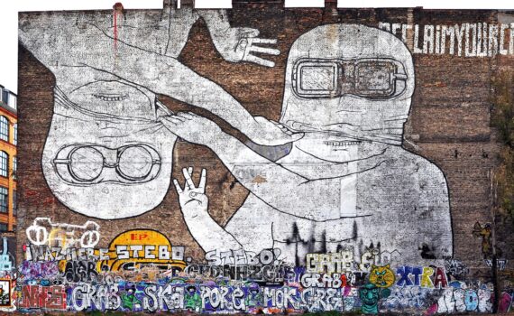 Große Wandtapete mit zwei Comicfiguren mit Schutzbrillen, von denen eine aufrecht und die andere auf dem Kopf steht, vor einer Backsteinmauer, die im unteren Bereich mit bunten Graffiti-Tags übersät ist. (KI generierte Beschreibung)