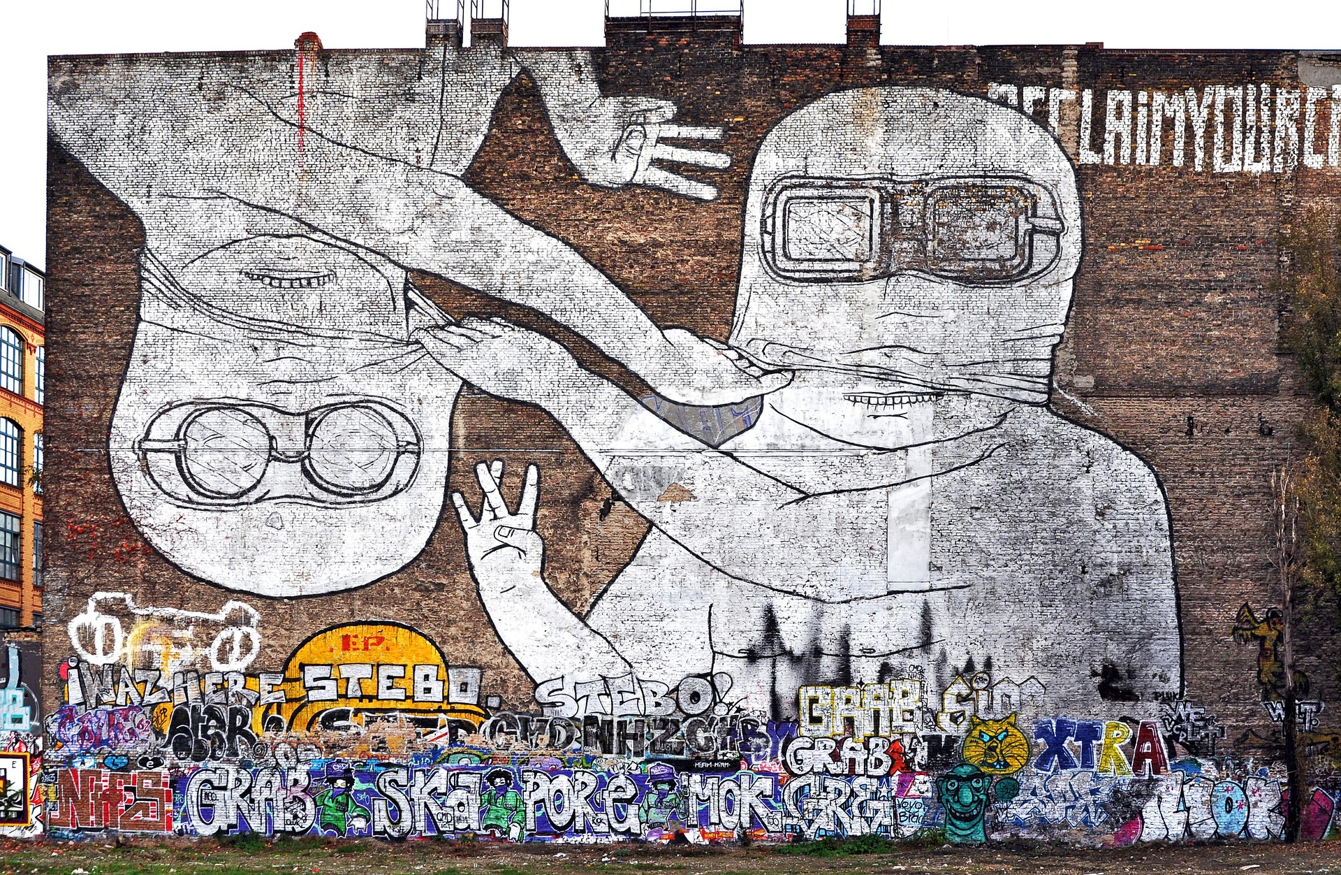 Große Wandtapete mit zwei Comicfiguren mit Schutzbrillen, von denen eine aufrecht und die andere auf dem Kopf steht, vor einer Backsteinmauer, die im unteren Bereich mit bunten Graffiti-Tags übersät ist. (KI generierte Beschreibung)