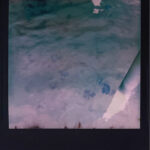Ein Polaroidfoto, das eine leicht unscharfe Unterwasserszene mit mehreren Quallen und einem diagonal verlaufenden weißen Pfahl oder Rohr zeigt. (KI generierte Beschreibung)