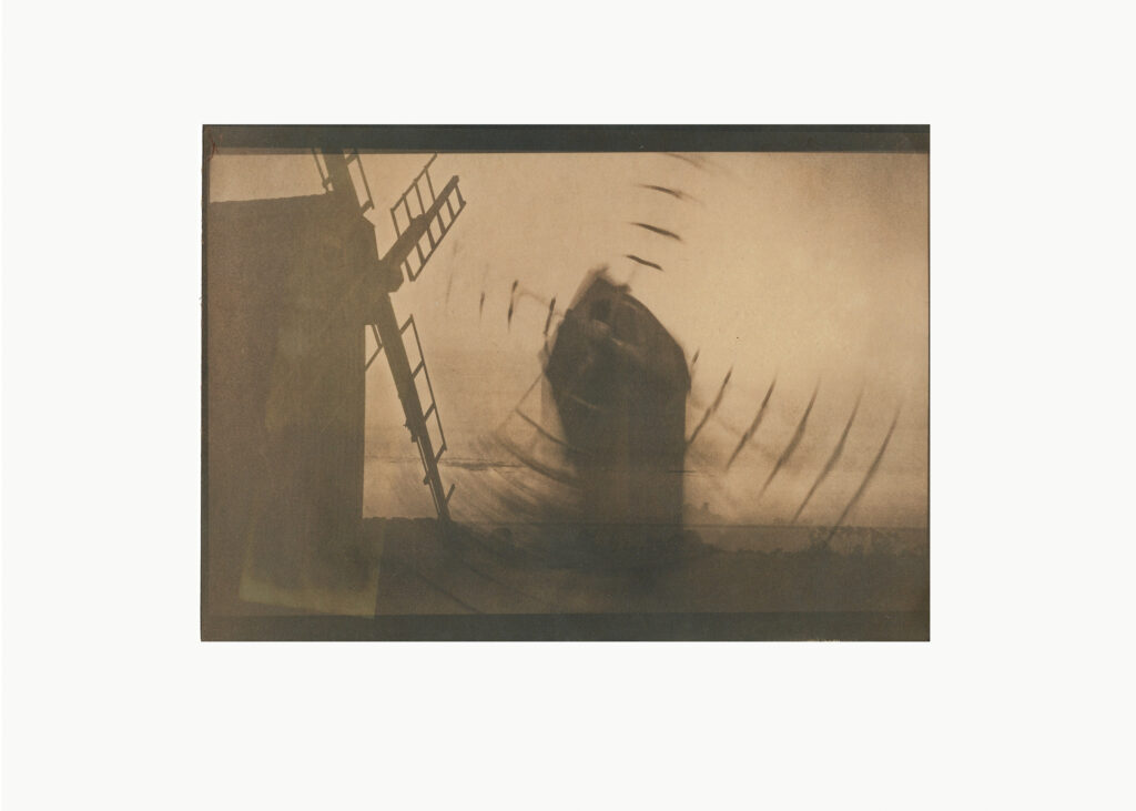 Ein sepiafarbenes Foto einer Windmühle mit verschwommenen, sich drehenden Flügeln und einer schwachen, abstrakten Form in der Mitte, die einen Eindruck von Bewegung vermittelt. (KI generierte Beschreibung)