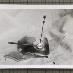 Schwarz-Weiß-Foto einer zerbrochenen Oldtimer-Kühlerfigur mit Uhrenmotiv, die auf einer Stoffunterlage liegt und von Glasscherben umgeben ist. (KI generierte Beschreibung)
