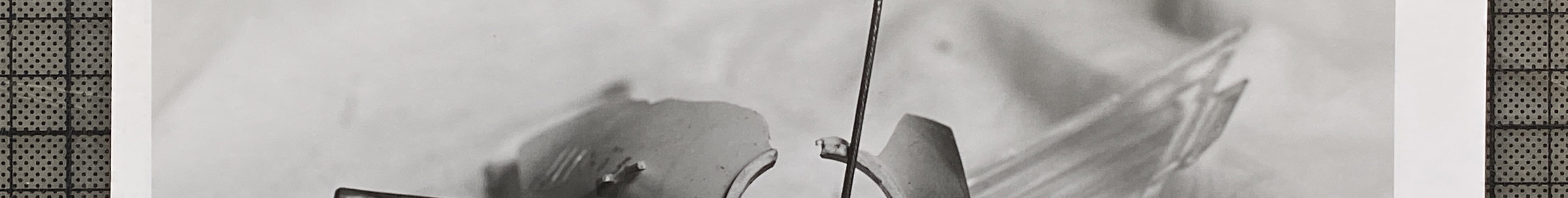 Schwarz-Weiß-Foto einer zerbrochenen Oldtimer-Kühlerfigur mit Uhrenmotiv, die auf einer Stoffunterlage liegt und von Glasscherben umgeben ist. (KI generierte Beschreibung)