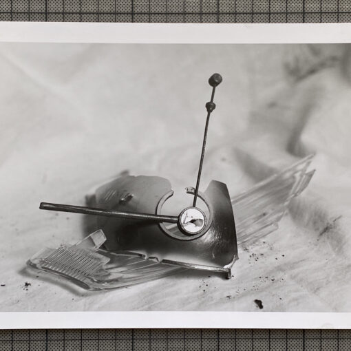 Schwarz-Weiß-Foto einer zerbrochenen Oldtimer-Kühlerfigur mit Uhrenmotiv, die auf einer Stoffunterlage liegt und von Glasscherben umgeben ist. (KI generierte Beschreibung)