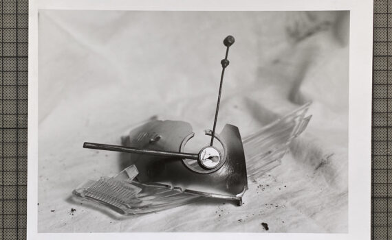 Schwarz-Weiß-Foto einer zerbrochenen Oldtimer-Kühlerfigur mit Uhrenmotiv, die auf einer Stoffunterlage liegt und von Glasscherben umgeben ist. (KI generierte Beschreibung)